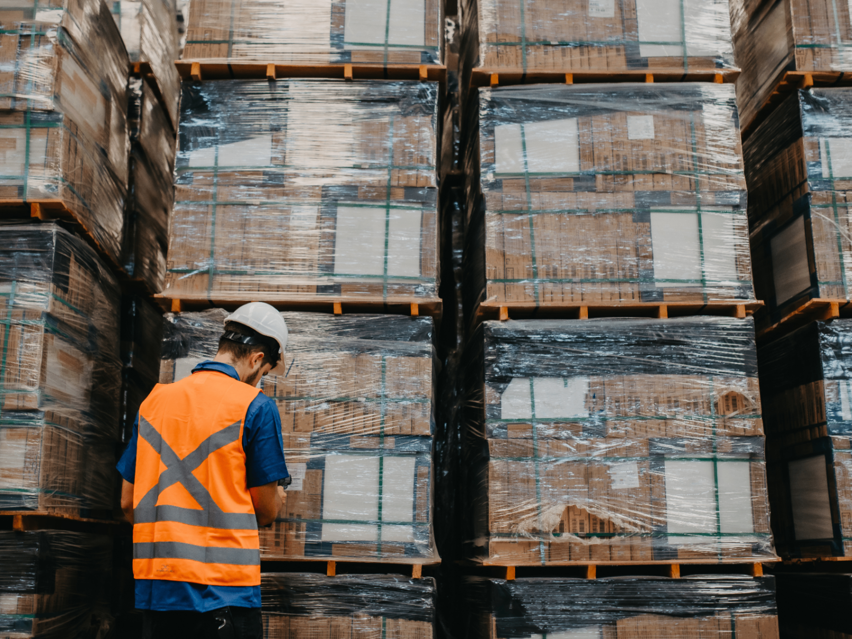 Warehouse worker in a high-visibility vest inspecting stacked, wrapped pallets in an equipment distribution facility
